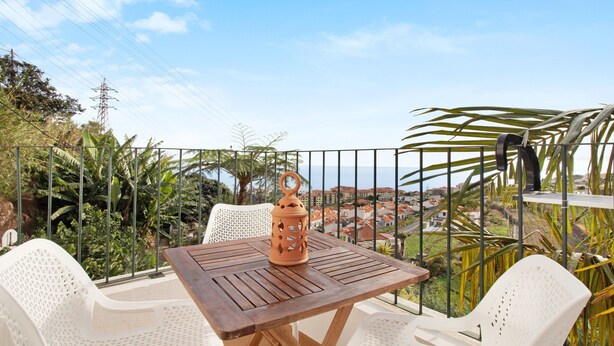 A balcony with a table and chairs, a decorative lantern, and a view of a coastal town. - Attic in 'Casa Amarela' with Sea & Mountain View, Wi-Fi, Garden, Parking & Garden (Caniço)