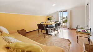 A room with a bed, a dining table with chairs, a small table with a vase, and a view of the outdoors. - Attic in 'Casa Amarela' with Sea & Mountain View, Wi-Fi, Garden, Parking & Garden (Caniço)