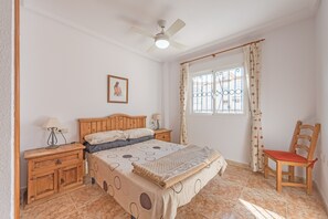 A bedroom with a bed, wooden nightstands, a chair, a ceiling fan, a window with curtains, and a wall-mounted picture.