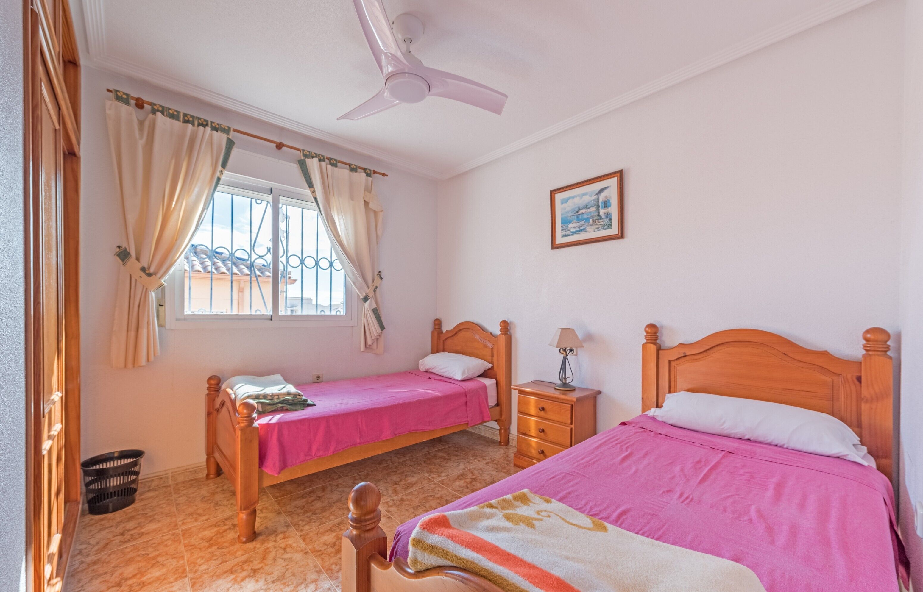 A bedroom with two beds, a ceiling fan, a window with curtains, a nightstand, and a wall-mounted picture.