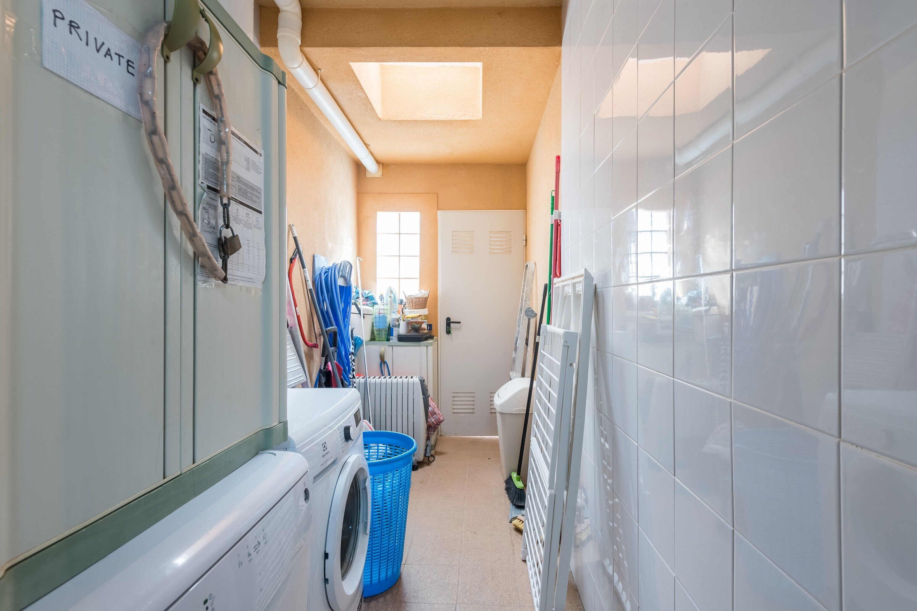 A narrow laundry room with a washing machine, a dryer, a blue laundry basket, and a radiator.
