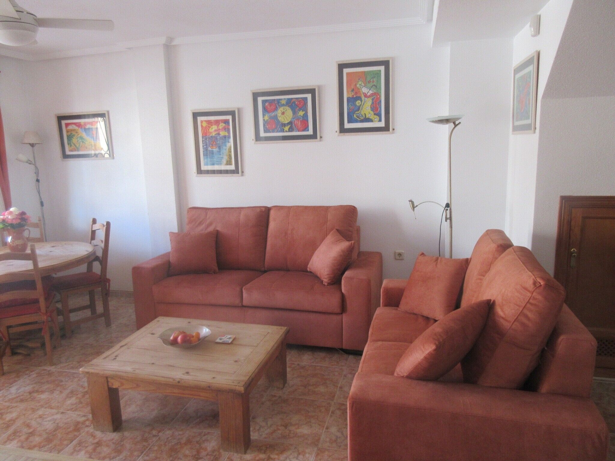A living room with a sofa, a coffee table, and a dining area with chairs. There are framed pictures on the wall.
