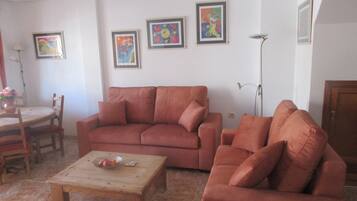A living room with a sofa, a coffee table, and a dining area with chairs. There are framed pictures on the wall.