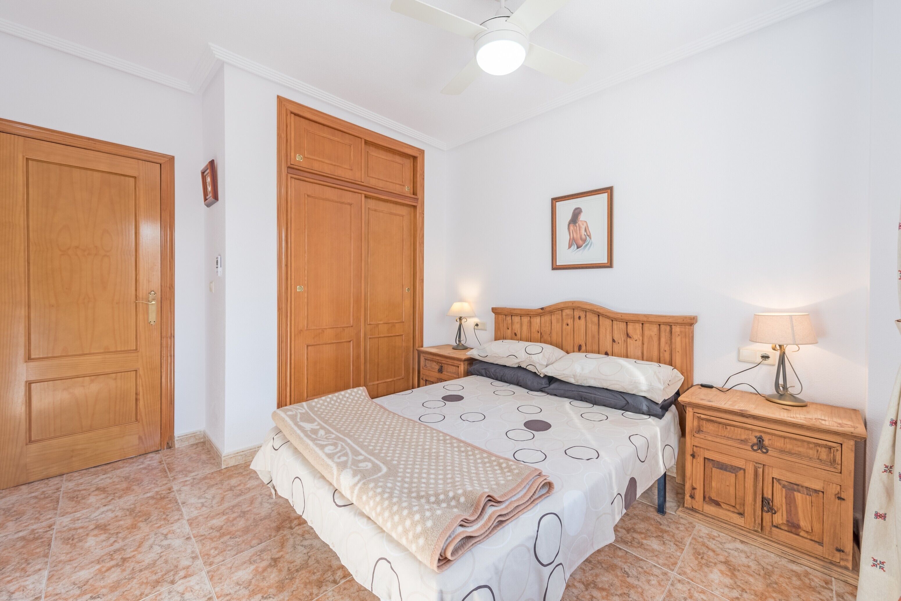 Une chambre à coucher comprenant un lit, une armoire en bois, des tables de chevet et un ventilateur de plafond.