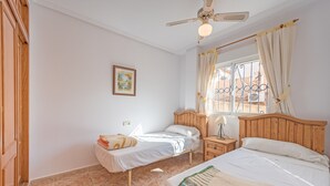 Een slaapkamer met twee eenpersoonsbedden, een houten hoofdbord, een plafondventilator, een raam met gordijnen en een klein nachtkastje met een lamp.