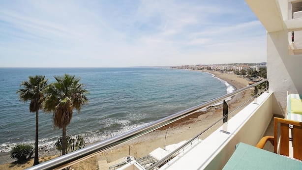 A balcony with a view of the sea and beach, a railing, and a table with a chair.