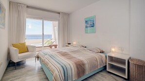 A bedroom with a bed, a chair, a bedside table, a lamp, a window with curtains, and a wall-mounted picture.