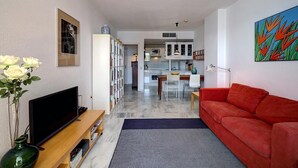 Un soggiorno con un divano rosso, una TV su un mobile in legno, una libreria e una cucina sullo sfondo.