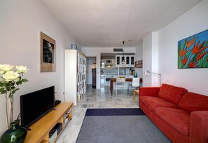 Een woonkamer met een rode bank, een televisie op een houten dressoir, een boekenkast en een keuken op de achtergrond.
