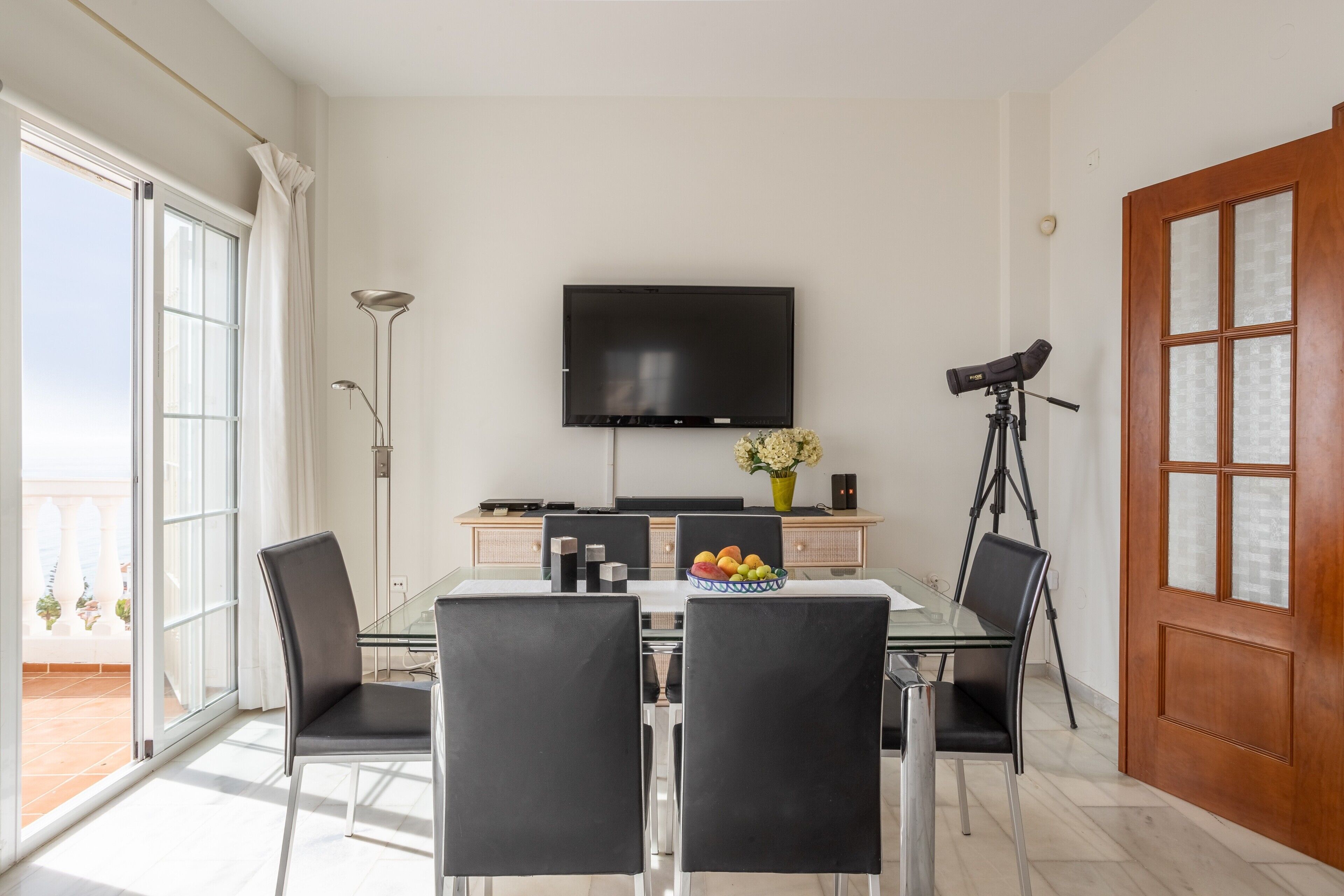 Un comedor moderno con una mesa de vidrio, sillas negras, un televisor de pantalla plana, un trípode y un bol de fruta.