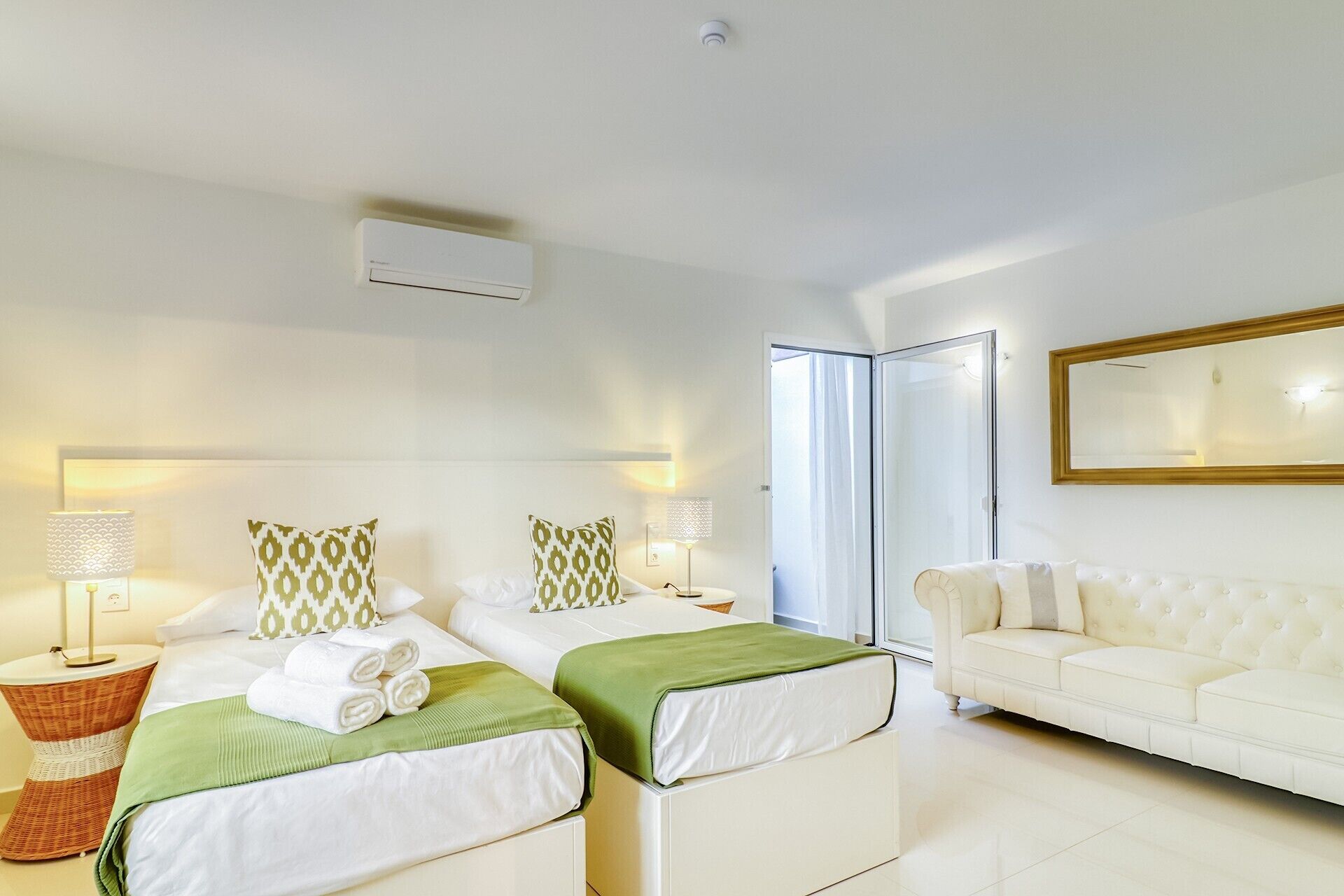 A hotel room with two beds, a sofa, a mirror, and a ceiling fan.
