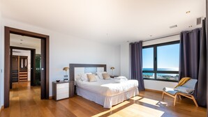 Ein modernes Schlafzimmer mit einem großen Bett, einem Sessel, einem Nachttisch und Blick auf den Ozean.