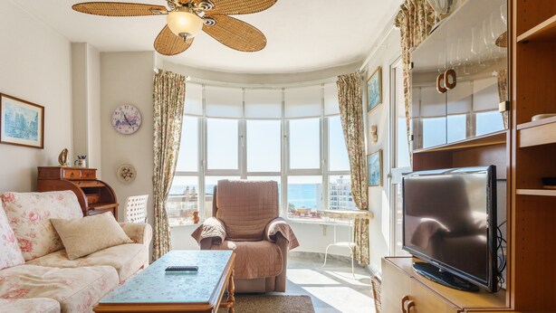 Una sala de estar con sofá, sillón, mesa de centro, televisor y ventanas amplias con cortinas.