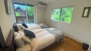 A bedroom with two beds, a window with a view of greenery, an air conditioning unit, and a wooden floor. - Coopers House Hakuba (Hakuba)