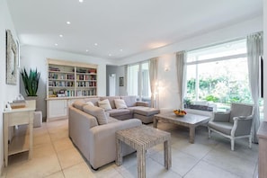 Ein modernes Wohnzimmer mit einem Ecksofa, einem Couchtisch, einem Bücherregal und einem großen Fenster mit Vorhängen.