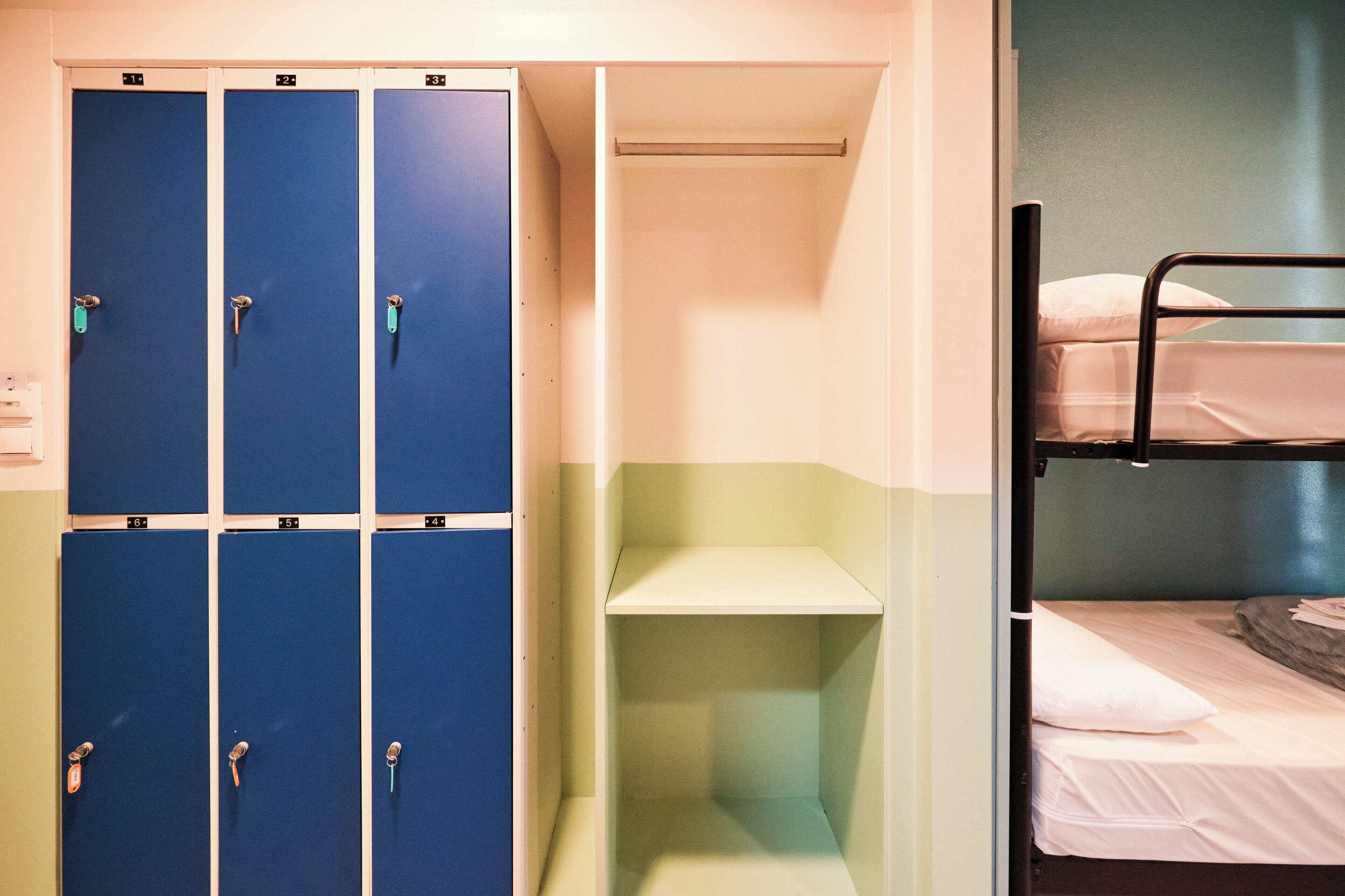 A dormitory room with bunk beds, a closet with blue lockers, and a small shelf unit.