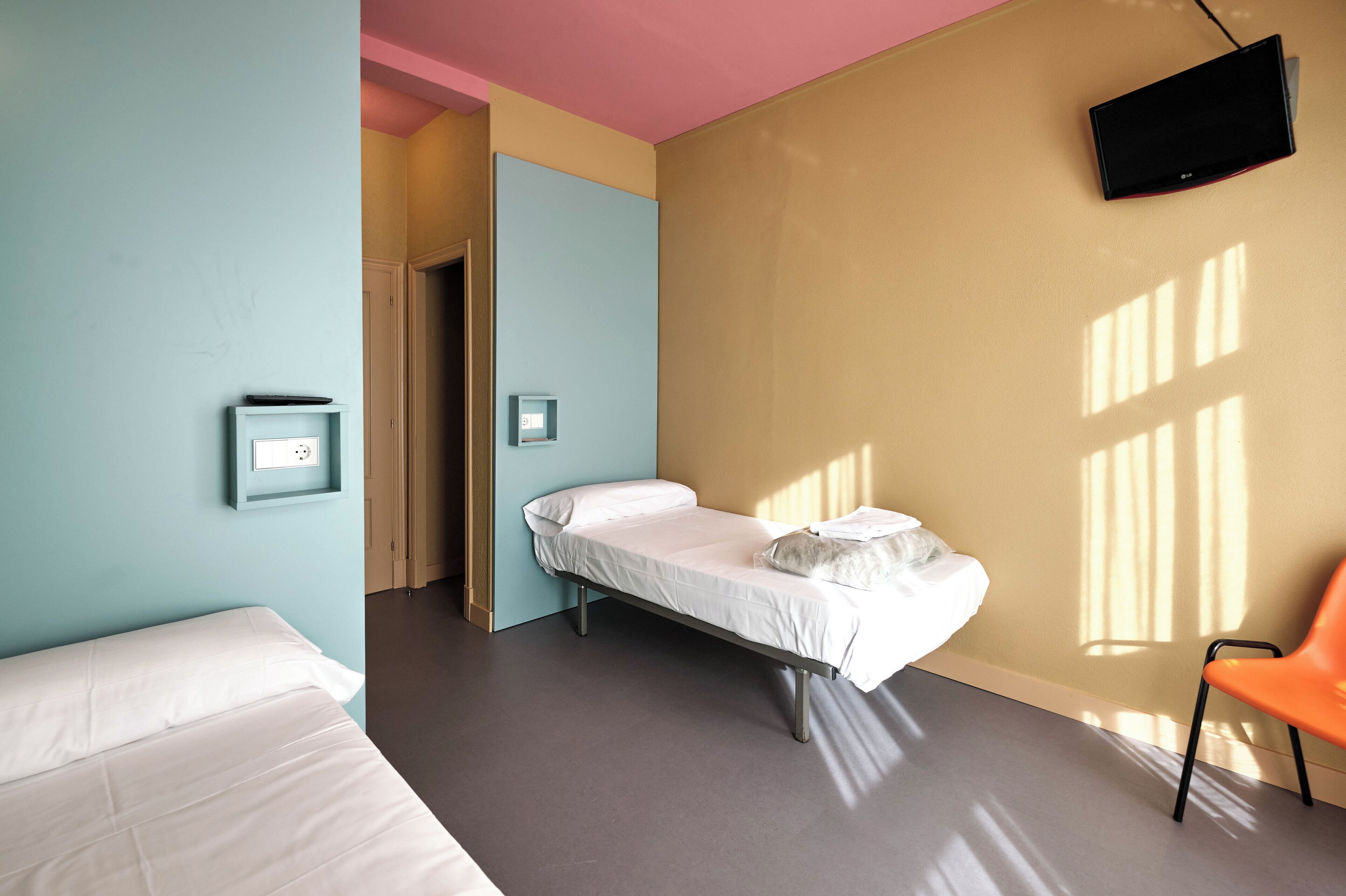 Una cama individual con colchón y almohadas blancas, una mesita de noche con lámpara, un televisor montado en la pared y una ventana que proyecta sombras sobre la pared.