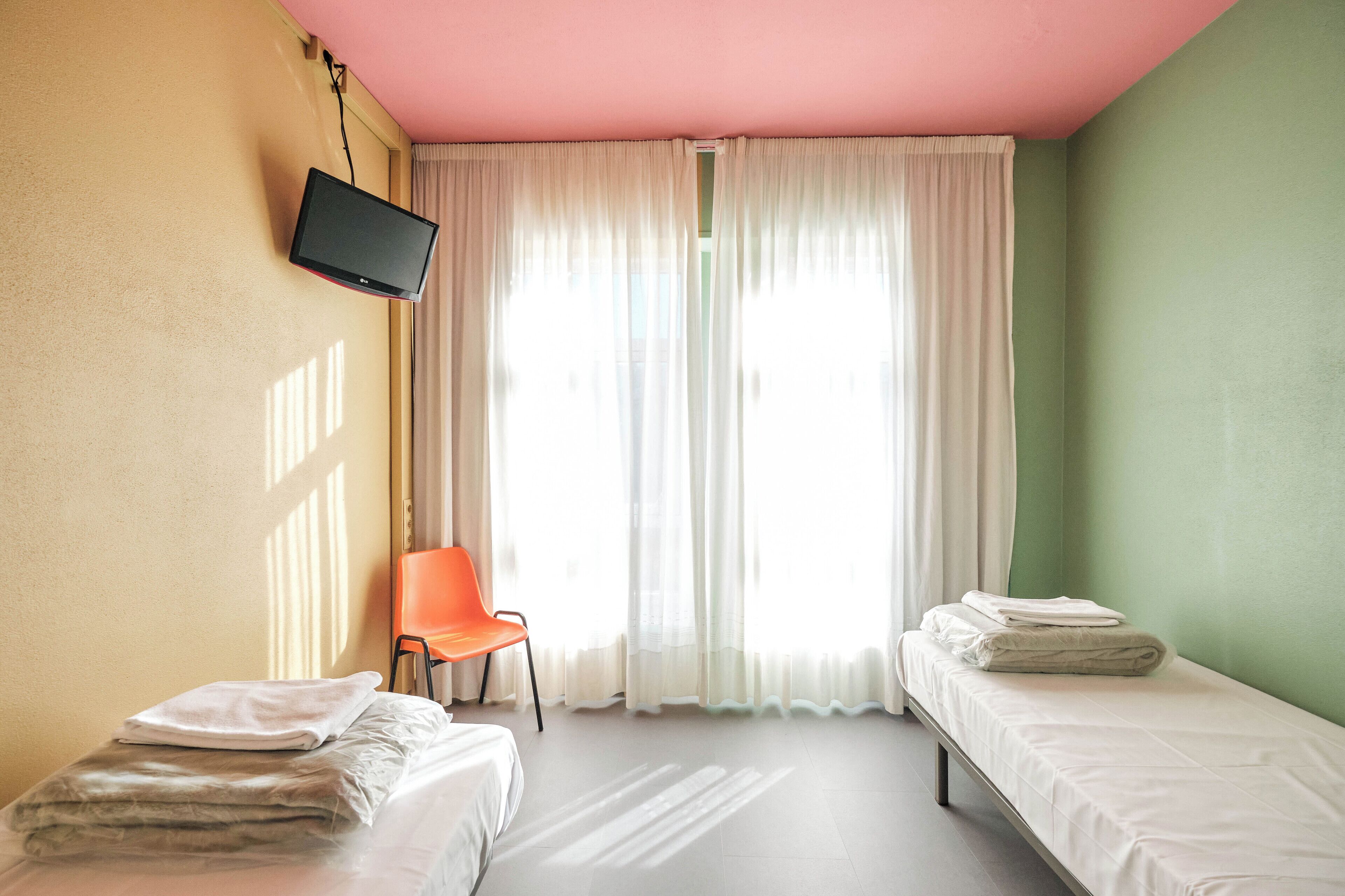 Una habitación con dos camas, un televisor montado en la pared, una silla naranja y una ventana con cortinas traslúcidas.