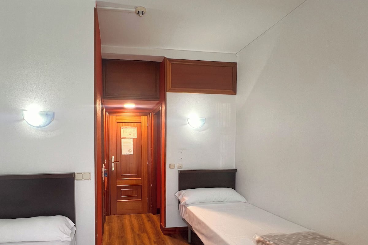 A small, compact room with two single beds, a wooden floor, and a door leading to another room.