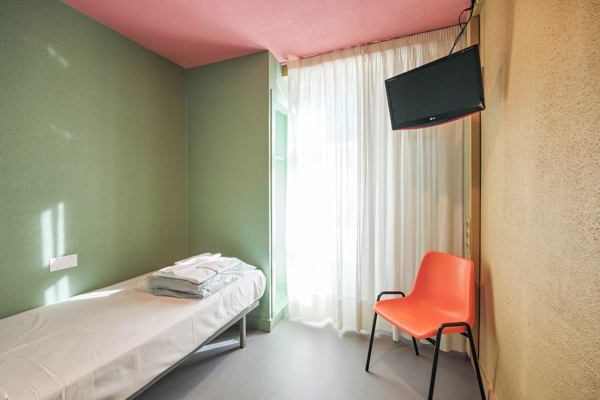 A single-bed room with a television on the wall, a chair, and a window with curtains.