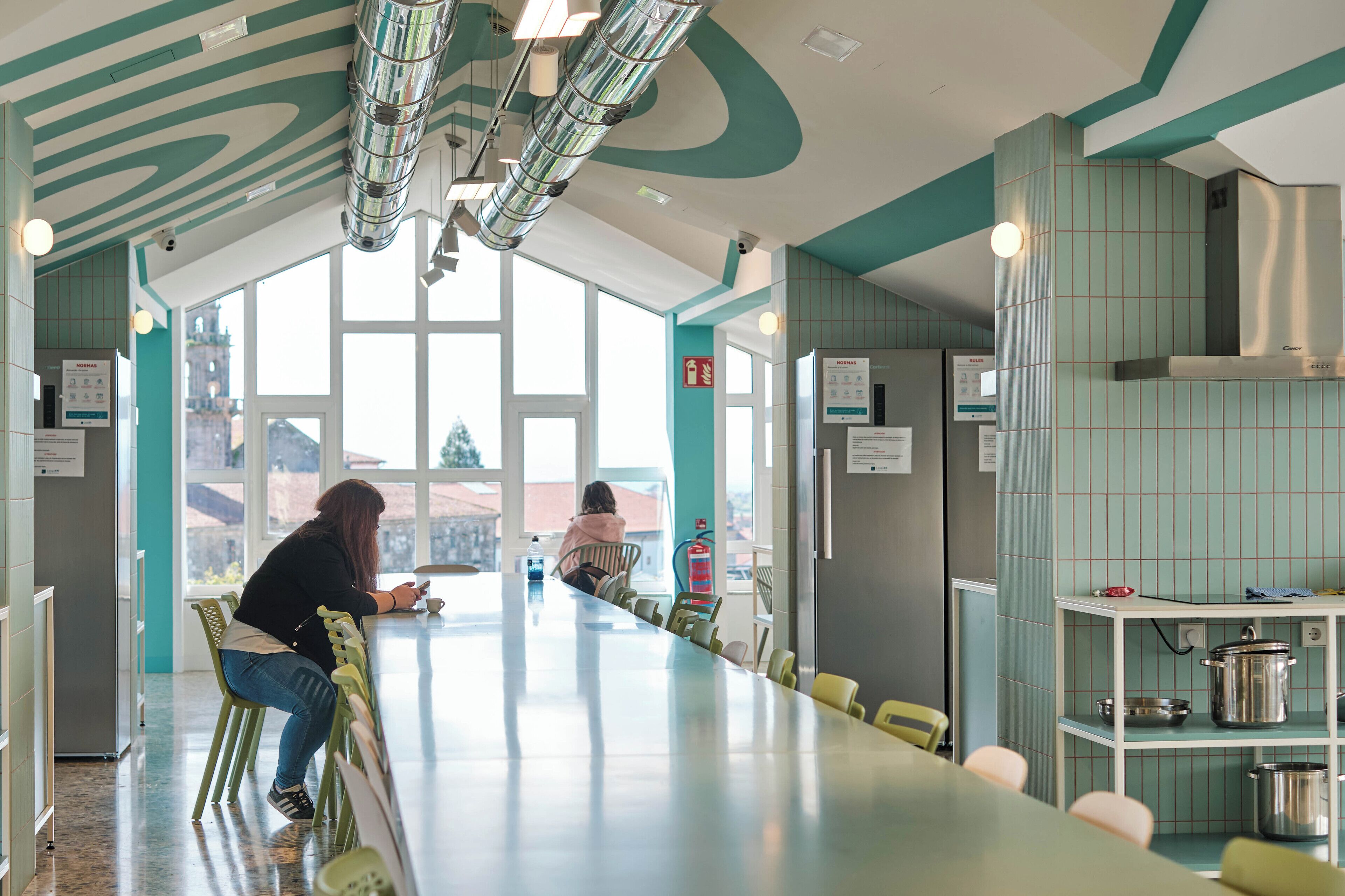 A long, narrow dining area with a high ceiling and large windows. A woman is seated at a table, using her phone. The walls are tiled in a light color, and there are several chairs and tables arranged along the length of the room.