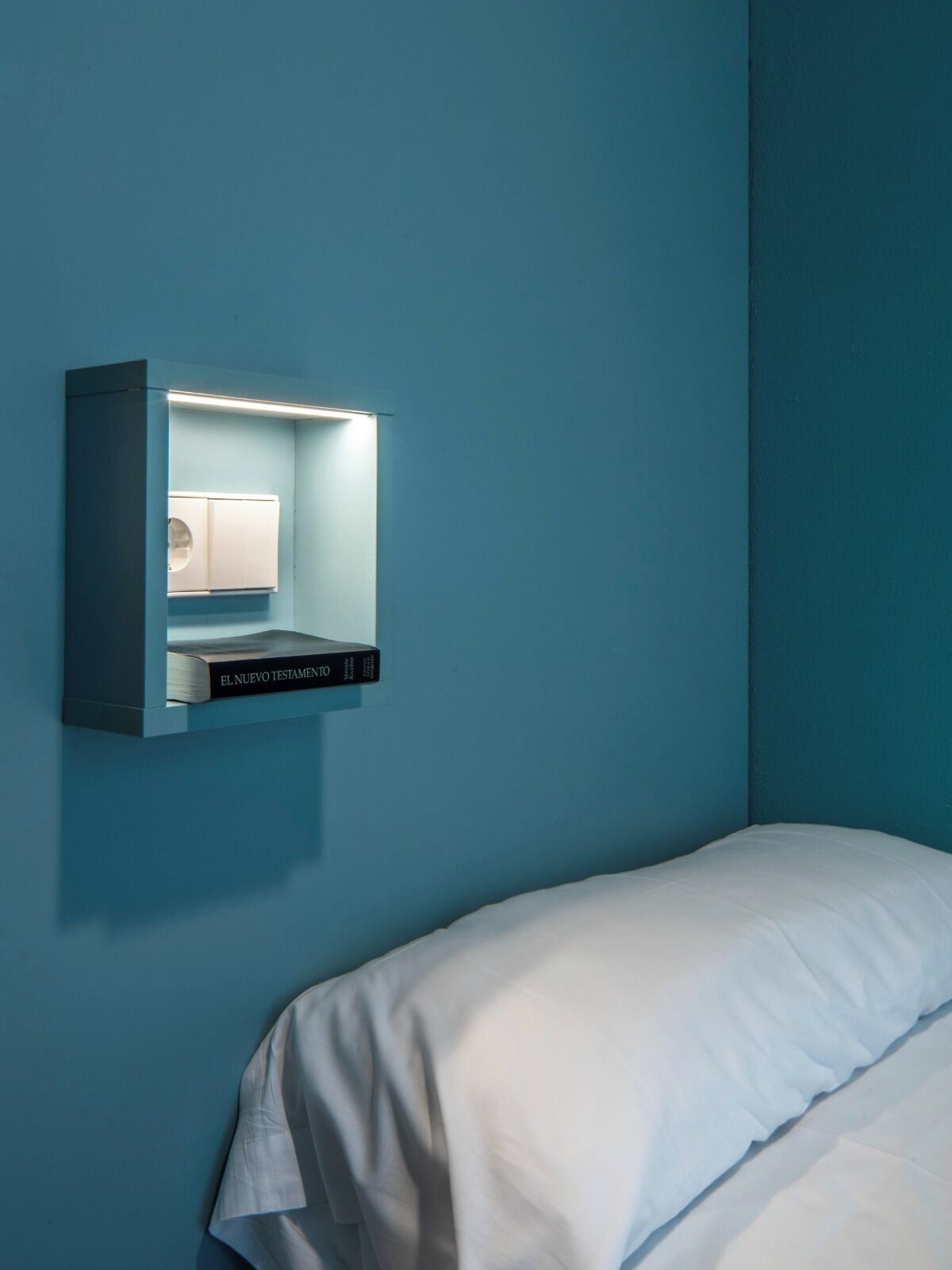 Une chambre avec un lit simple, une lampe de lecture fixée au mur et un livre intitulé « EL NUEVO TESTAMENTO ».