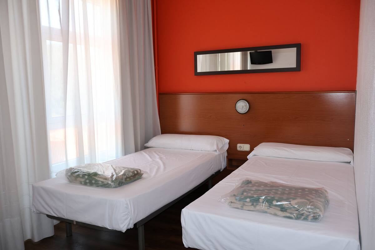 A hotel room with two single beds, a wooden headboard, a wall-mounted mirror, and a window with sheer curtains.
