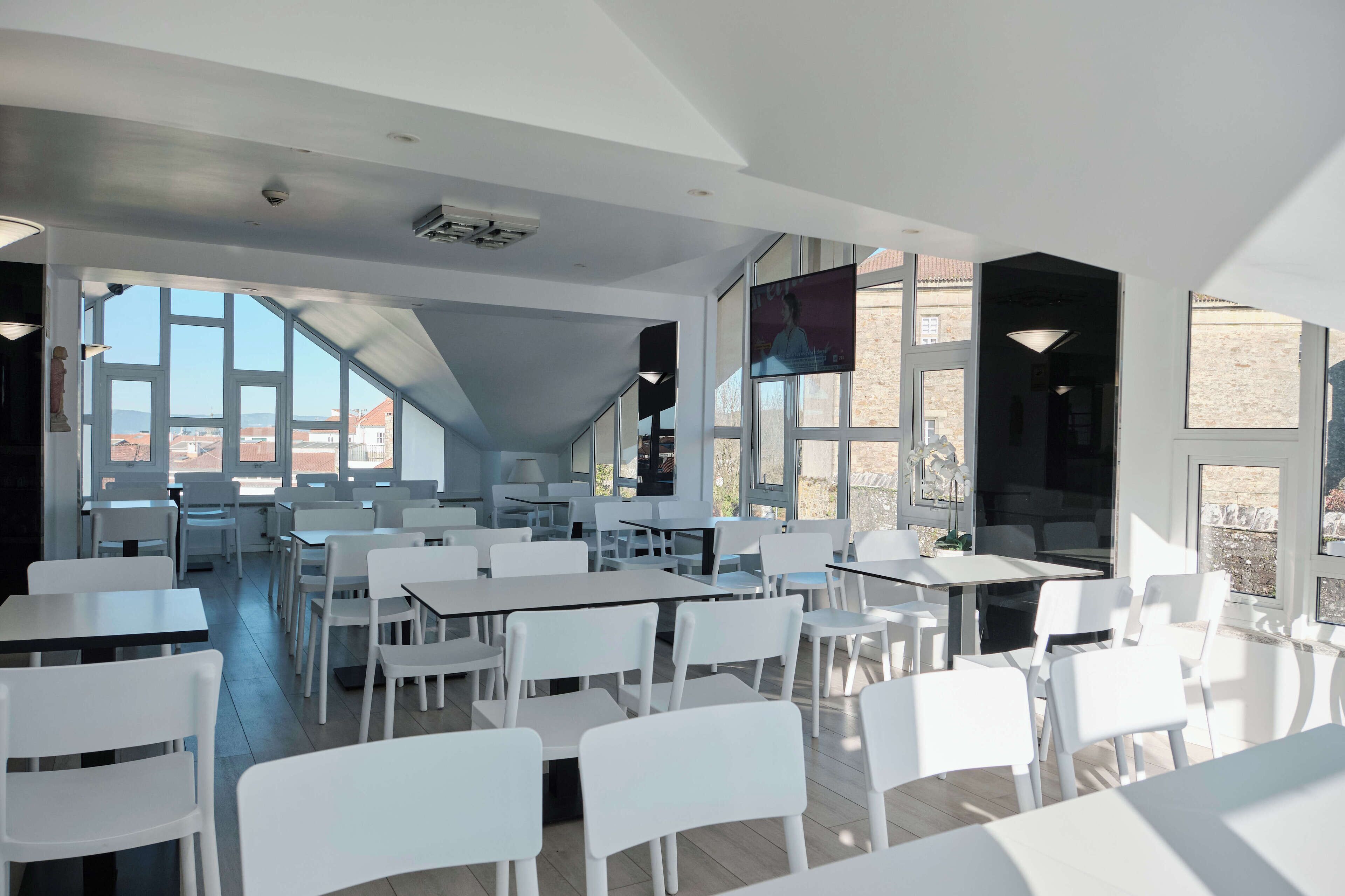 A spacious, modern dining area with white tables and chairs, large windows offering views of the city, and a flat-screen TV mounted on the wall.