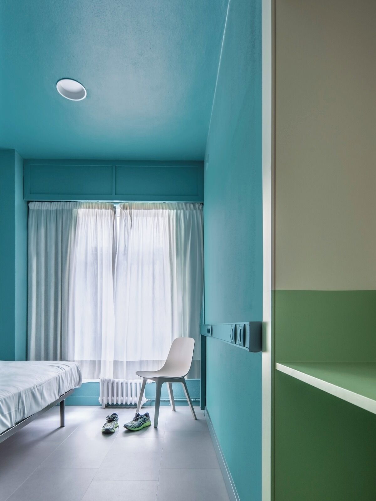 Une pièce aux murs de couleur turquoise, avec un lit blanc, une chaise blanche et une paire de baskets posées sur le sol.