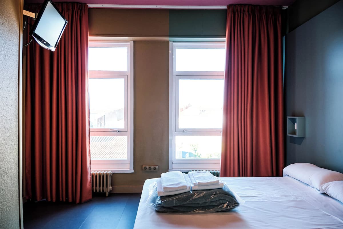 A hotel room with two windows, a bed with neatly folded white towels, a radiator, and a wall-mounted TV.