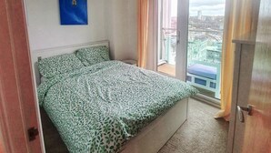 Un dormitorio con cama, una lámpara en el techo, vista de la ciudad a través de una ventana y una puerta.