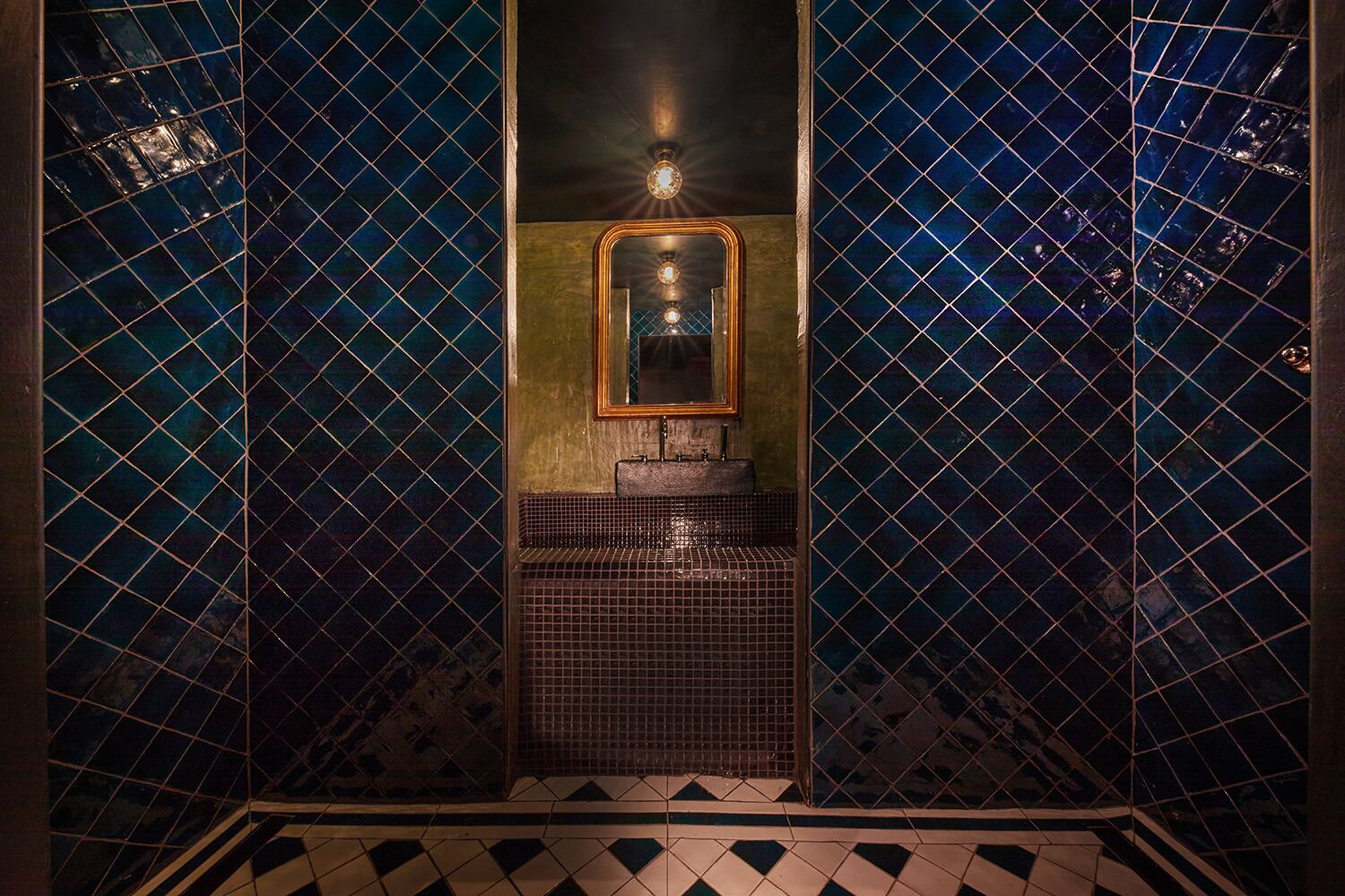 A bathroom with a dark blue mosaic tile wall, a gold-framed mirror, and a sink with a tiled base.
