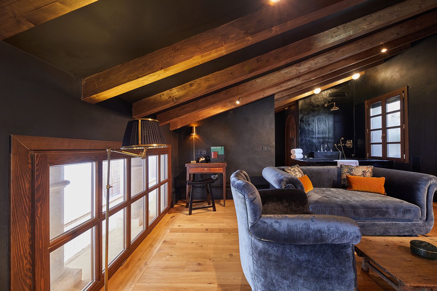 Um quarto estilo loft aconchegante com vigas de madeira aparentes, uma janela grande com persianas, um sofá azul macio e uma pequena mesa de madeira com uma luminária.