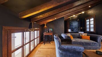 Um quarto estilo loft aconchegante com vigas de madeira aparentes, uma janela grande com persianas, um sofá azul macio e uma pequena mesa de madeira com uma luminária.