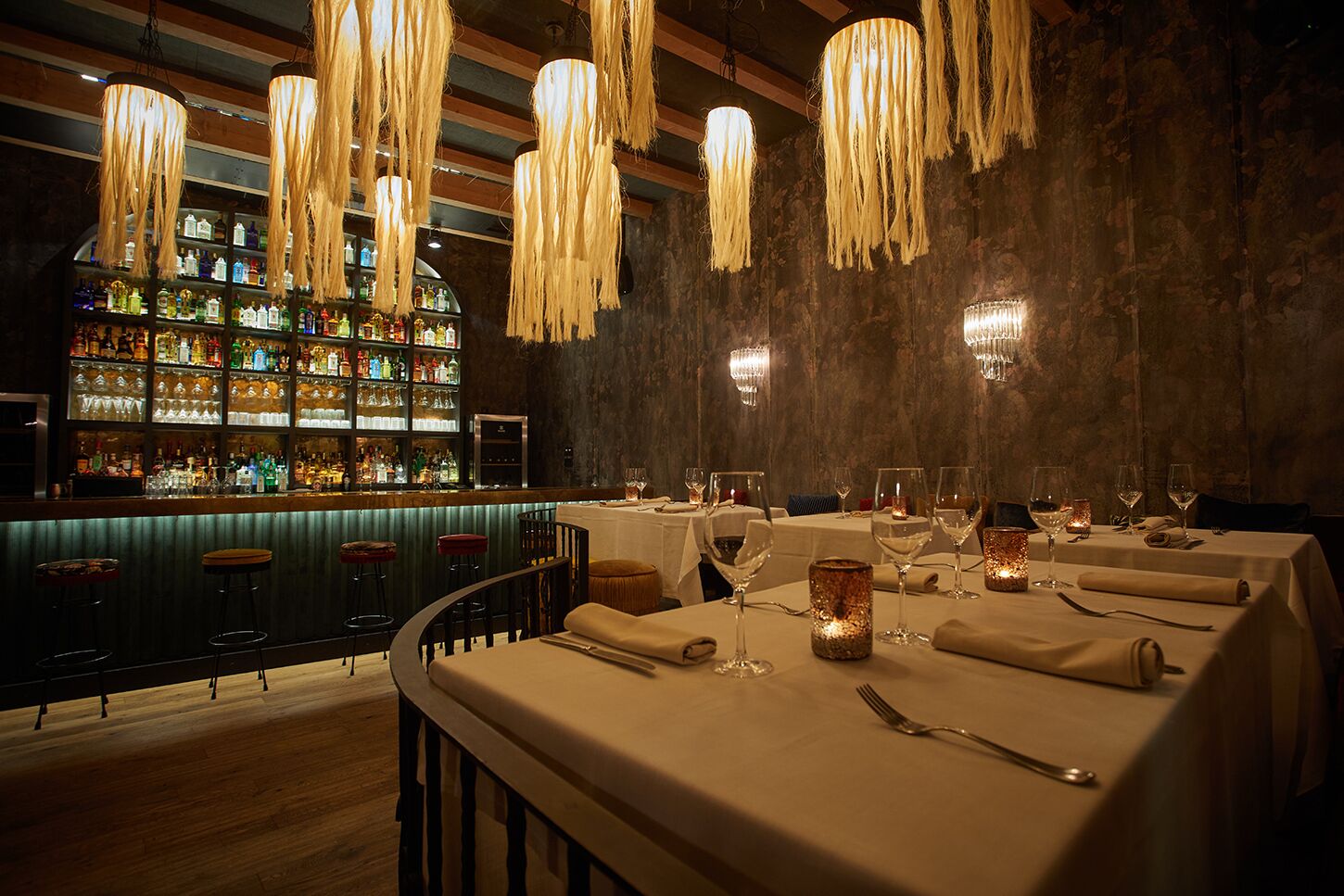 Um restaurante com várias mesas preparadas para refeições, área de bar com diversos tipos de garrafas e luminárias pendentes.