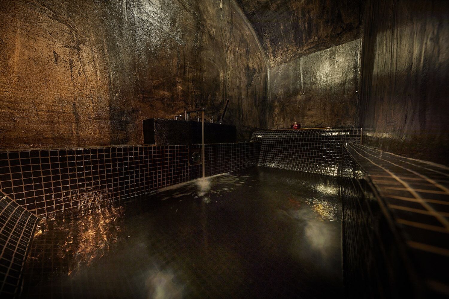 A dimly lit, tiled indoor pool with a single faucet and a small container on the wall.