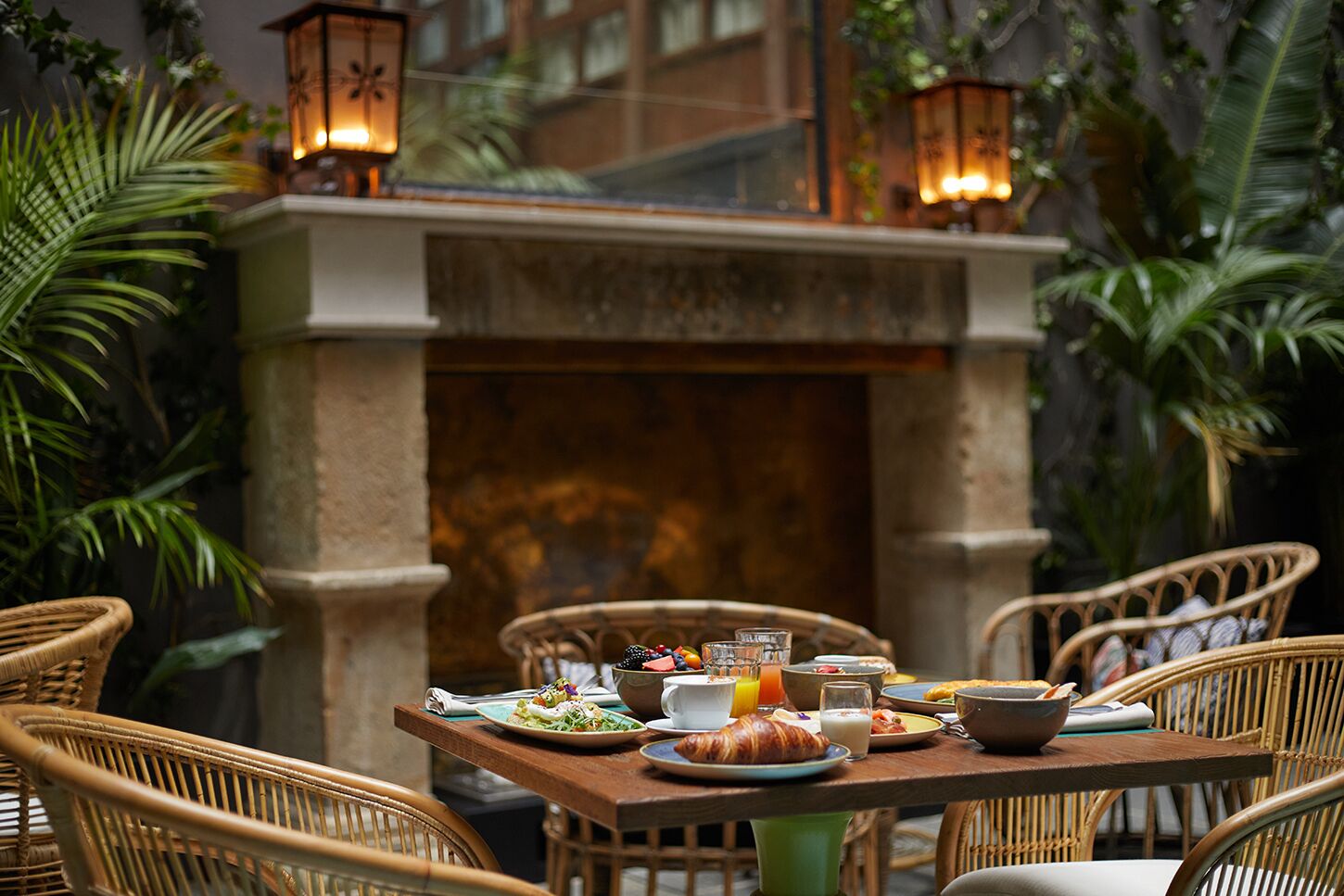 An outdoor dining area with wicker chairs and a table set with breakfast items, surrounded by lush greenery and a stone fireplace.