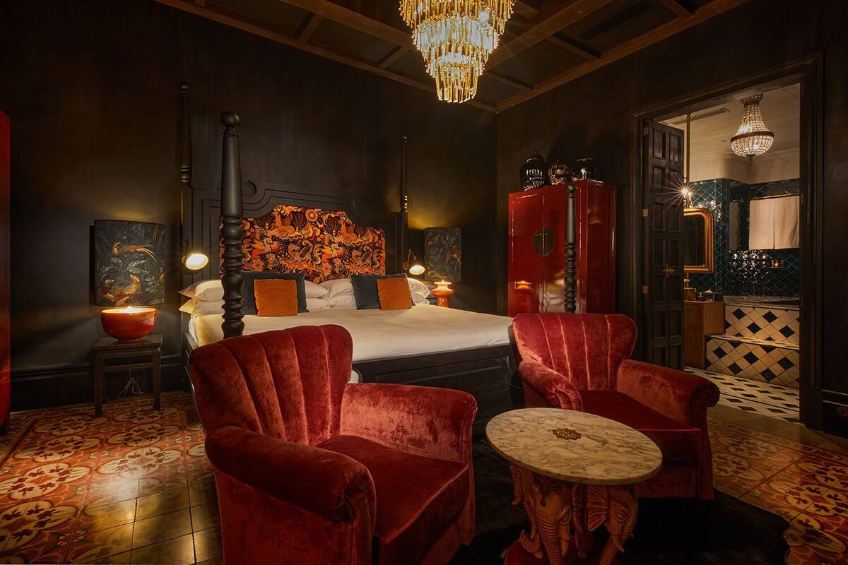 Une chambre à coucher luxueuse avec un grand lit, des fauteuils en velours rouge, une table d’appoint en marbre et un lustre.