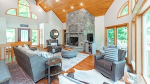 A spacious living room with a stone fireplace, wooden ceiling, and large windows offering views of the outdoors.