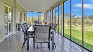 A covered outdoor area with a table and chairs, large windows, and a view of a grassy field.