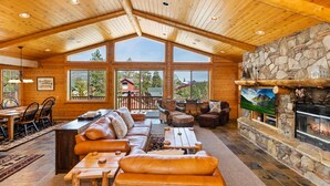 A spacious lodge with a high wooden ceiling, large windows offering views of the surrounding landscape, a stone fireplace, and a cozy seating area with leather sofas and a coffee table.