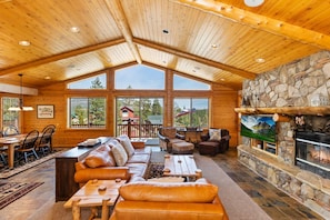 A spacious lodge with a high wooden ceiling, large windows offering views of the surrounding landscape, a stone fireplace, and a cozy seating area with leather sofas and a coffee table.