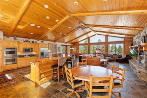 A spacious kitchen with wooden cabinets, a large island, and a dining area with a wooden table and chairs. - Rebel Ridge Retreat by Destination Big Bear : Gorgeous Mountain Views Backing to Nature Preserve in Eagle Mountain Estates! 2 Master Suites! Hot Tub! Pet Friendly! (Big Bear Lake)