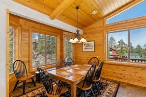 A dining area with a wooden table and chairs, large windows, and a ceiling with wooden beams. - Rebel Ridge Retreat by Destination Big Bear : Gorgeous Mountain Views Backing to Nature Preserve in Eagle Mountain Estates! 2 Master Suites! Hot Tub! Pet Friendly! (Big Bear Lake)