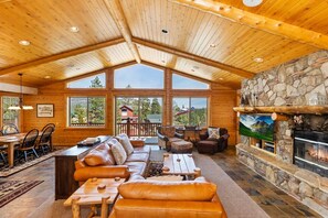 A spacious living area with a high ceiling, wooden beams, a stone fireplace, and large windows offering a view of the outdoors. - Rebel Ridge Retreat by Destination Big Bear : Gorgeous Mountain Views Backing to Nature Preserve in Eagle Mountain Estates! 2 Master Suites! Hot Tub! Pet Friendly! (Big Bear Lake)