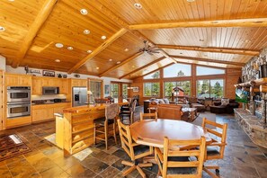 A spacious kitchen with wooden cabinetry, a central island, and a dining area with a large table and chairs. - Rebel Ridge Retreat by Destination Big Bear : Gorgeous Mountain Views Backing to Nature Preserve in Eagle Mountain Estates! 2 Master Suites! Hot Tub! Pet Friendly! (Big Bear Lake)