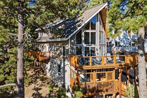 A two-story wooden house with a deck, surrounded by pine trees. - Window Wonderland by Destination Big Bear : Custom Luxury Built Near the Lake! Breathtaking Lake Views! Gameroom! Hot Tub! (Big Bear Lake)