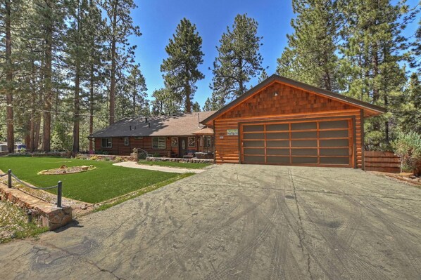 Cozy cabin with a garage, surrounded by a well-maintained lawn and tall pine trees, featuring a front porch with outdoor seating.