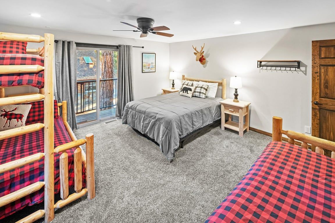 Ein Schlafzimmer mit Etagenbett, einem Einzelbett, einem Deckenventilator, einem Fenster mit Vorhängen, einem aufgehängten Hirschkopf und einer Holztür.