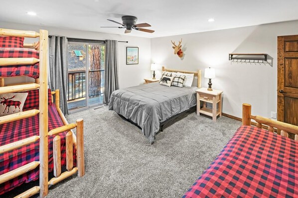 A bedroom with a bunk bed, a single bed, a ceiling fan, a window with curtains, a mounted deer head, and a wooden door.